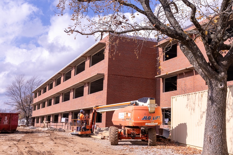 A three-story red brick building under construction with dirt in the foreground and cloudy sky in the background.