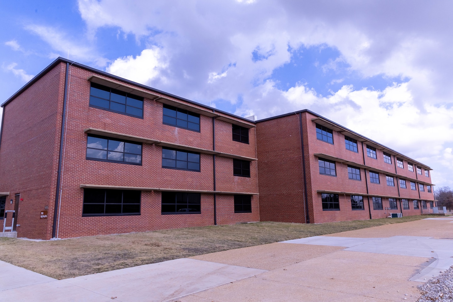 A red brick building with three stories and windows with grass and concrete in the foreground and blue sky with clouds in the background.