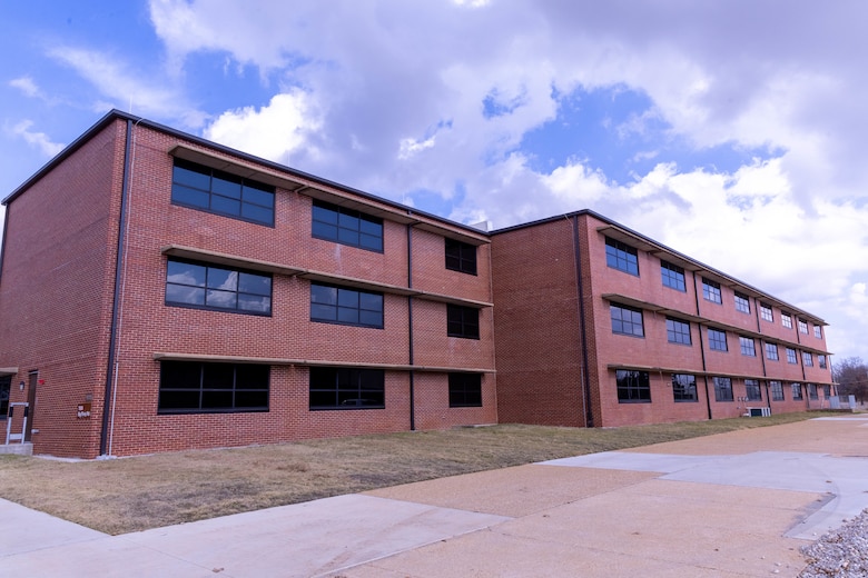 A red brick building with three stories and windows with grass and concrete in the foreground and blue sky with clouds in the background.