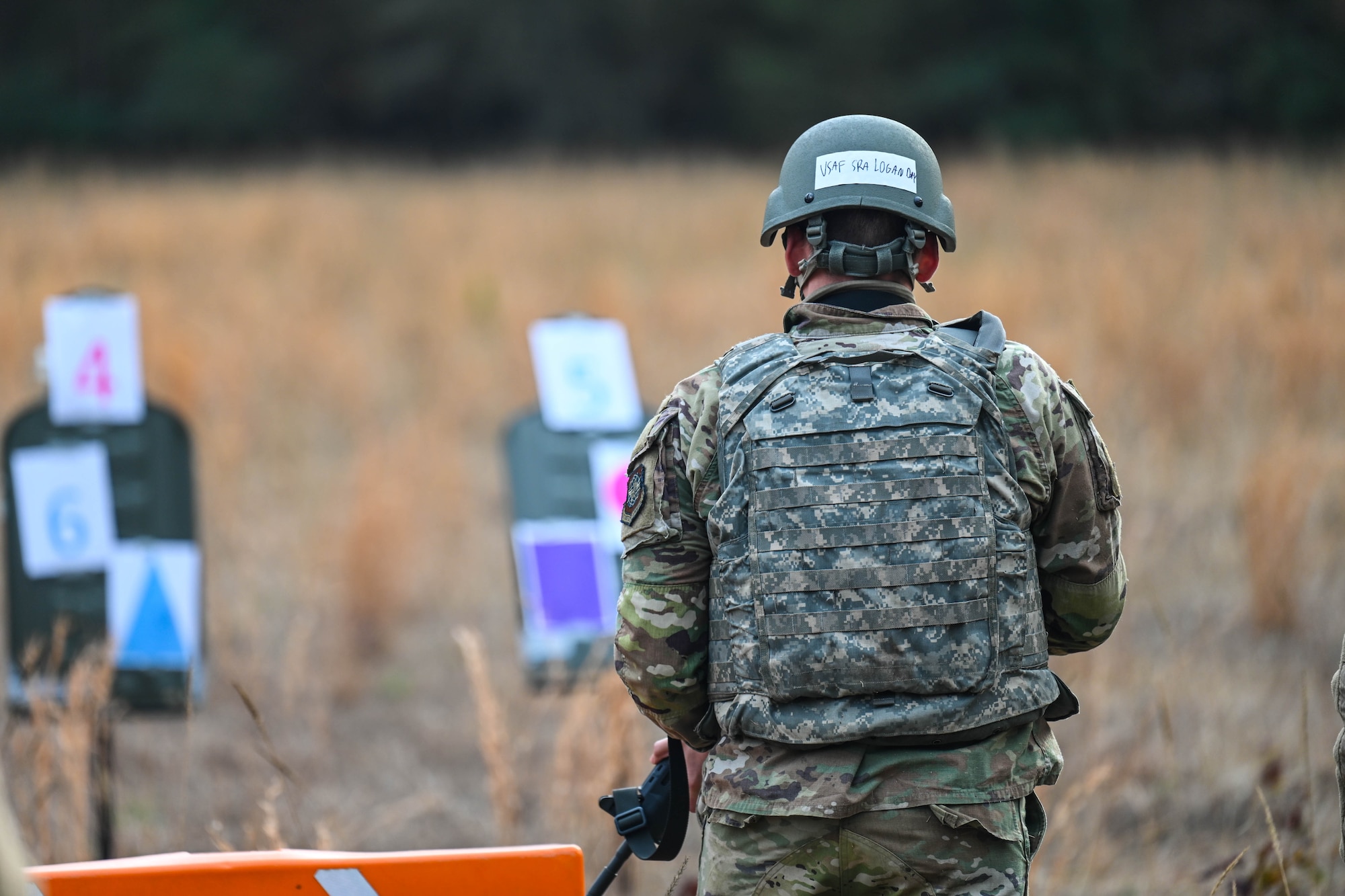 An Airman prepares to fire during a weapons training exercise. Targets are visible in the background with numbers and symbols.
