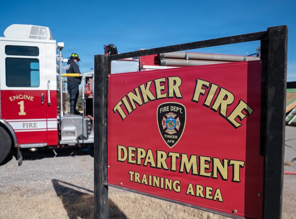 A red fire engine is visible in the background of a red and gold sign saying, "Tinker Fire Department Training Area."