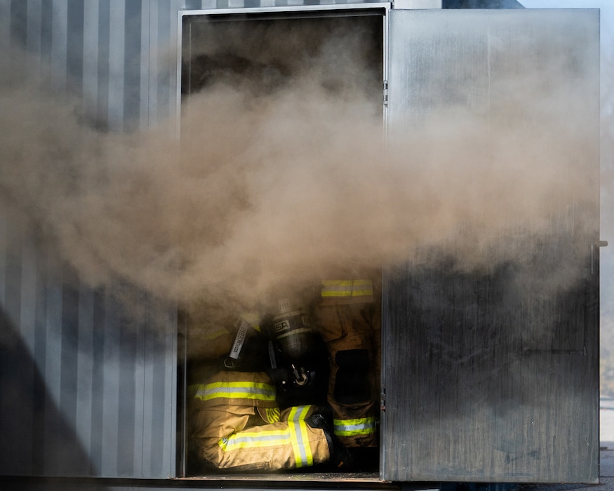 The lower half of a firefighter is visible kneeling in a doorway billowing with dark smoke.