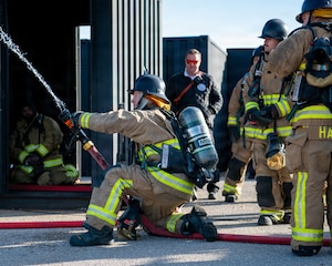 A firefighter kneels on the ground with a firehose in hand, shooting a stream of water .