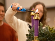 Close up of a woman lighting a purple candle with gold tips and gold base. There is a Christmas wreath underneath the candle.