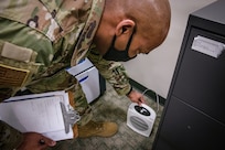 Close up of a military member bending down and inspecting a space heater. He is holding a clipboard and is touching the heater.