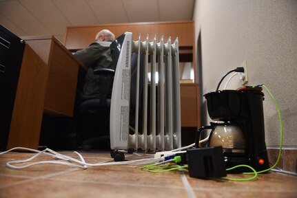 Man works at his desk. Space heater is shown close up and plugged into an outlet. Coffee maker is on the floor plugged into an extension cord.