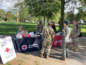 A woman stands at a table covered with snacks she is surrounded by men and women wearing military uniforms.