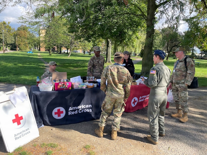 A woman stands at a table covered with snacks she is surrounded by men and women wearing military uniforms.