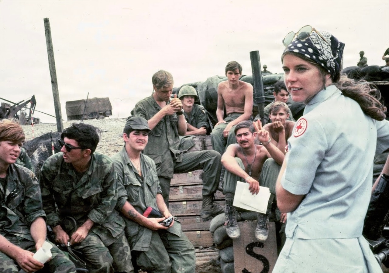 An American Red Cross volunteer stands to the right. She is surrounded by soldiers in various states of dress in this Vietnam War-era photo.