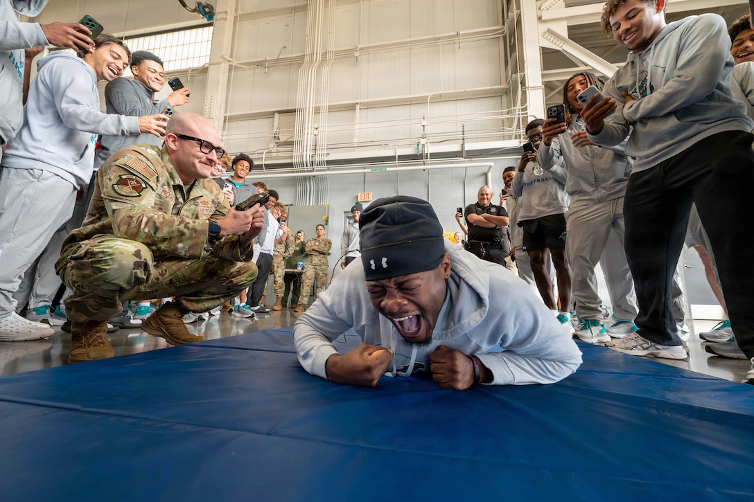 An airman shocks a screaming football player laying on a mat with a taser as other football players film with their phones.