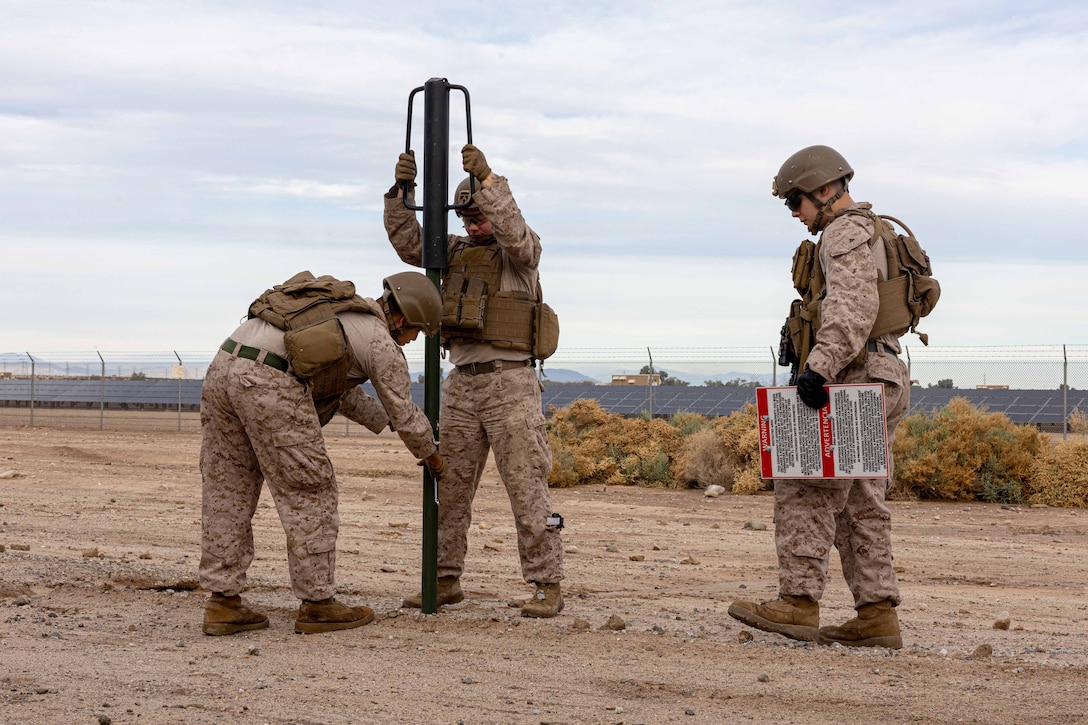 Marines put a sign post into the ground as a fellow service member watches in a desert-like area.