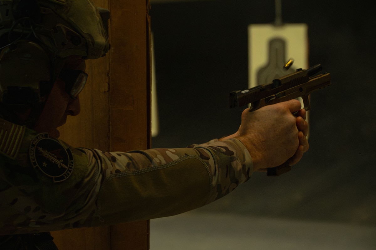 An Airman in tactical gear fires a pistol. The shell casing from the spent round is flying out of the chamber. A target is hanging in the background.