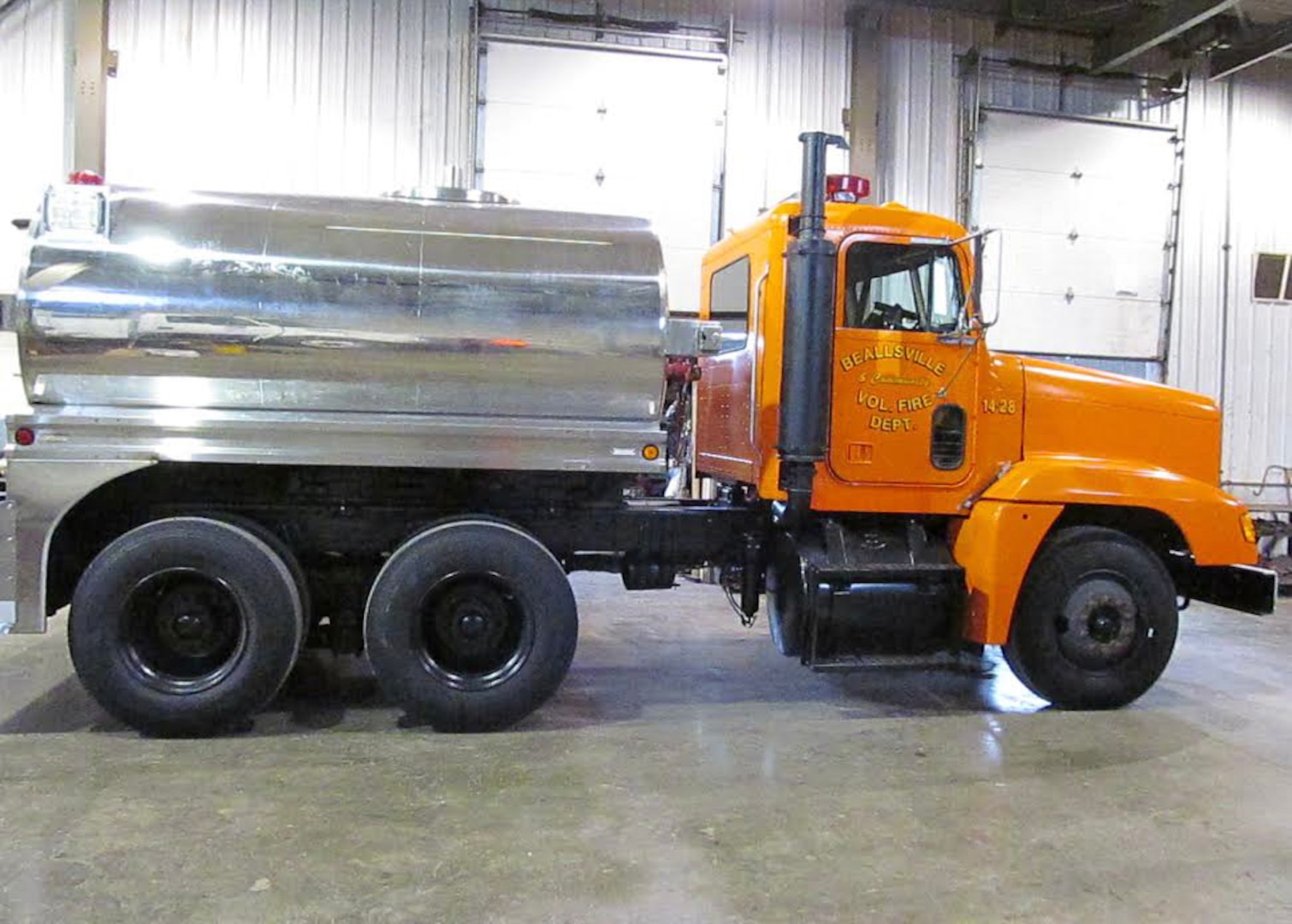 a former military truck with it's cab not painted orange and a large silver tank on the back sits inside a building.