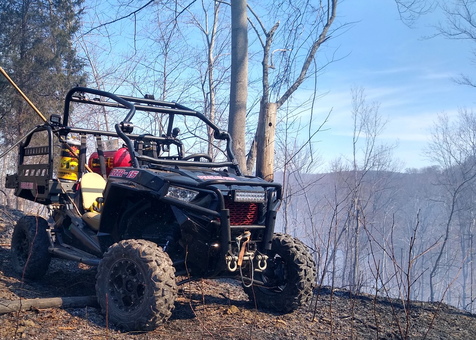 A small quad type vehicle sits on top of a ridge line with forest land in the distance background. The near ground looks as if a fire may have just taken place with a hint of smoke in the air.