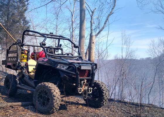A small quad type vehicle sits on top of a ridge line with forest land in the distance background. The near ground looks as if a fire may have just taken place with a hint of smoke in the air.