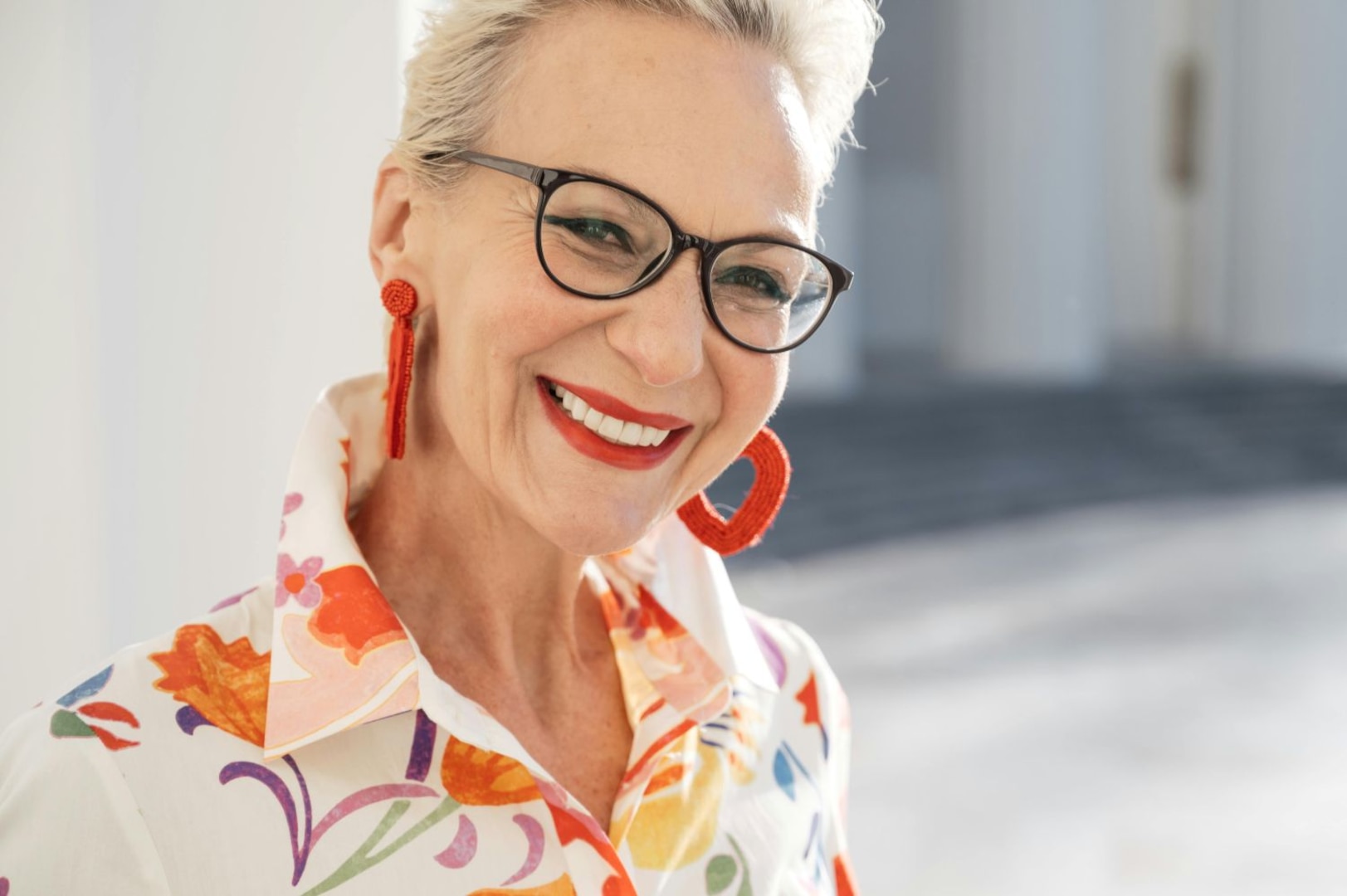 Elderly woman wearing floral shirt, earrings, and glasses smiles at the camera