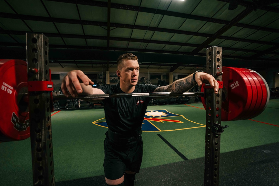 A soldier leans on a loaded squat rack in a dimly lit training facility.
