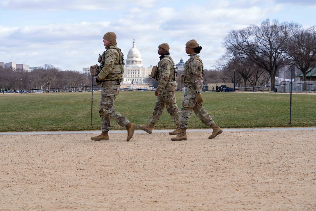 Three guardsmen walk on a dirt path, with the U.S. Capitol in the distance.