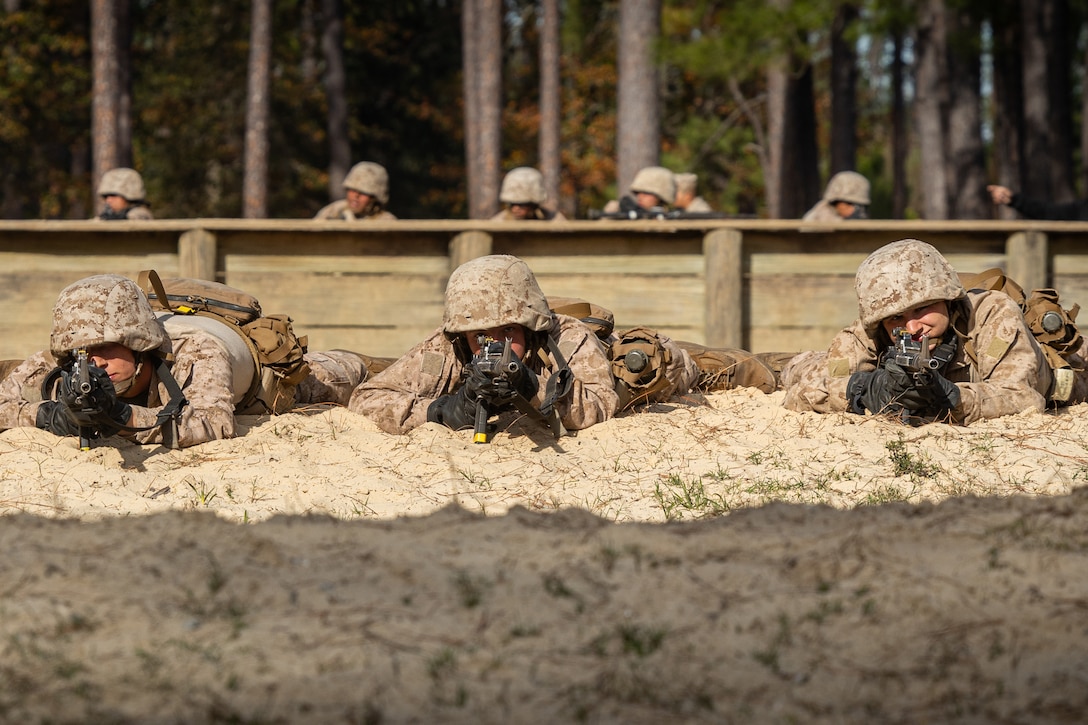 Three Marine Corps recruits lying in the dirt aim weapons as fellow service members work in the background.