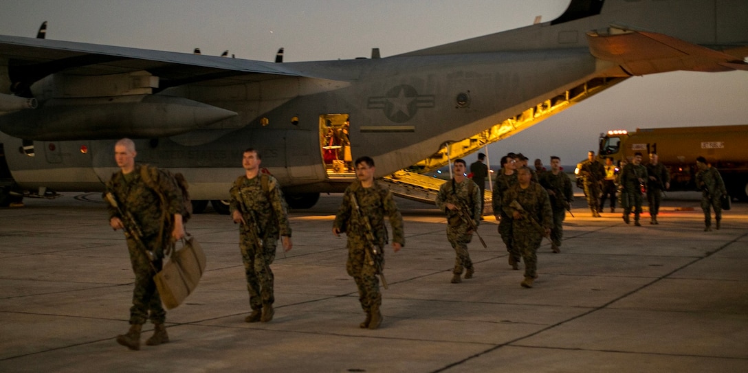 A line of military personnel in camouflage uniforms depart the back of an aircraft.