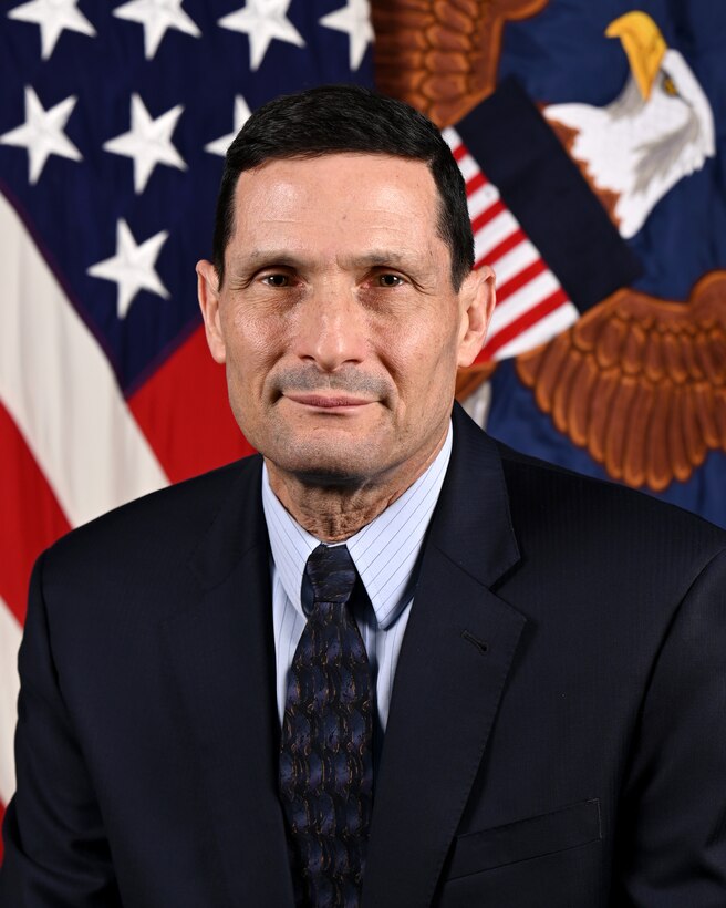 A man in a business suit poses in front of two flags.