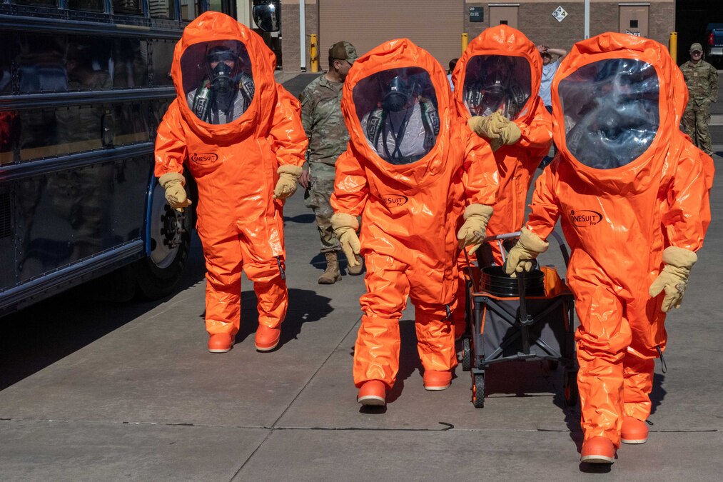 Four airmen walk fully covered in orange hazmat suits next to a black bus on pavement during the day.