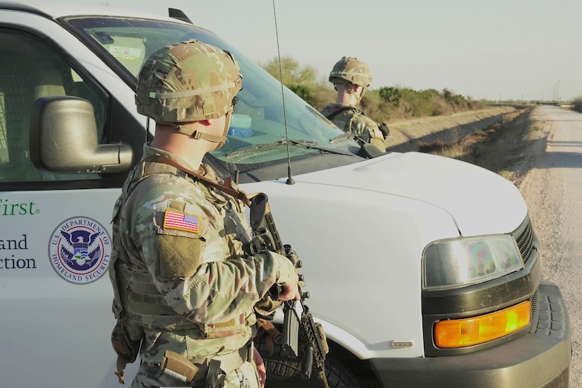 Two soldiers in camouflage uniforms are standing at the edge of a road near a van labeled 