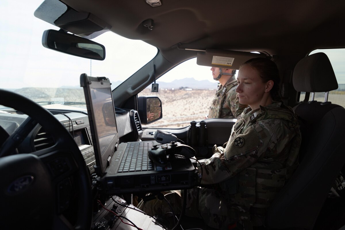 A female soldier in a camouflage uniform is sitting in the front seat of a passenger vehicle, observing a laptop computer screen.