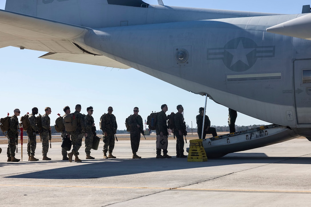 About 10 Marines stand in a line to board the open back of an aircraft parked on a flight line.