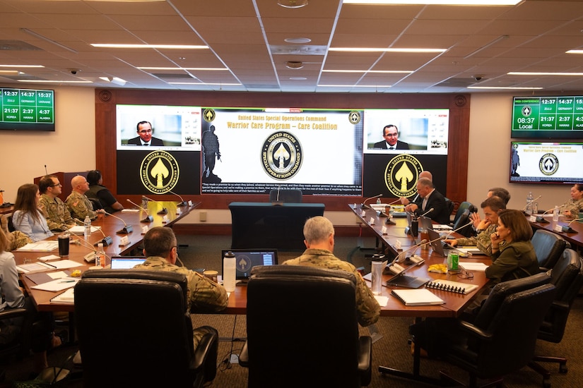 People in uniform sit at a U-shaped conference table with three screens in the background.