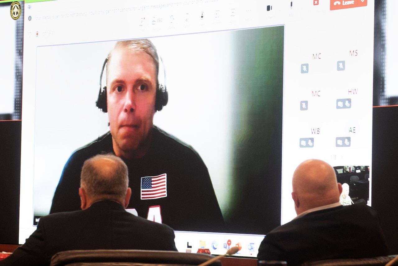 A man on a large projector screen speaks in a conference room.