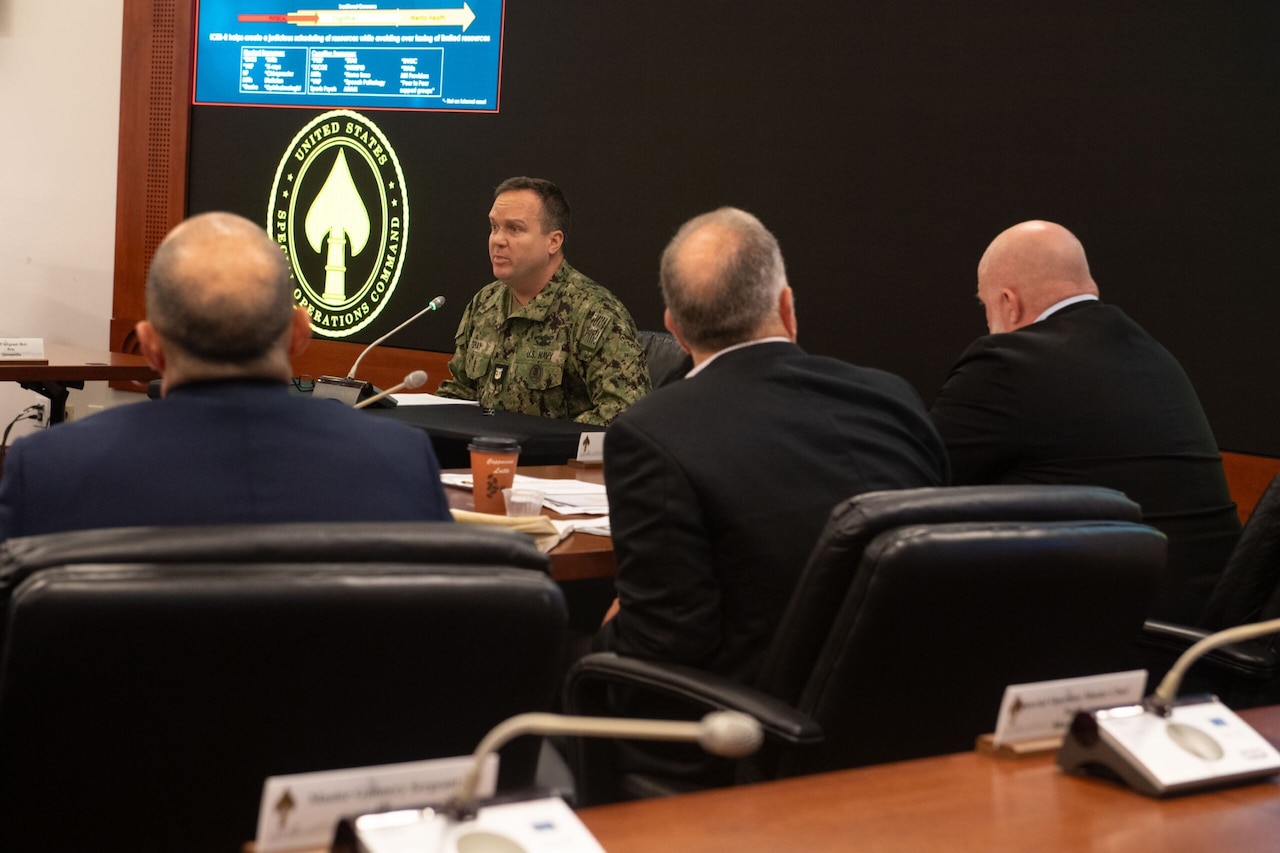 A man in uniform sits and speaks into a microphone while three audience members watch in the foreground.