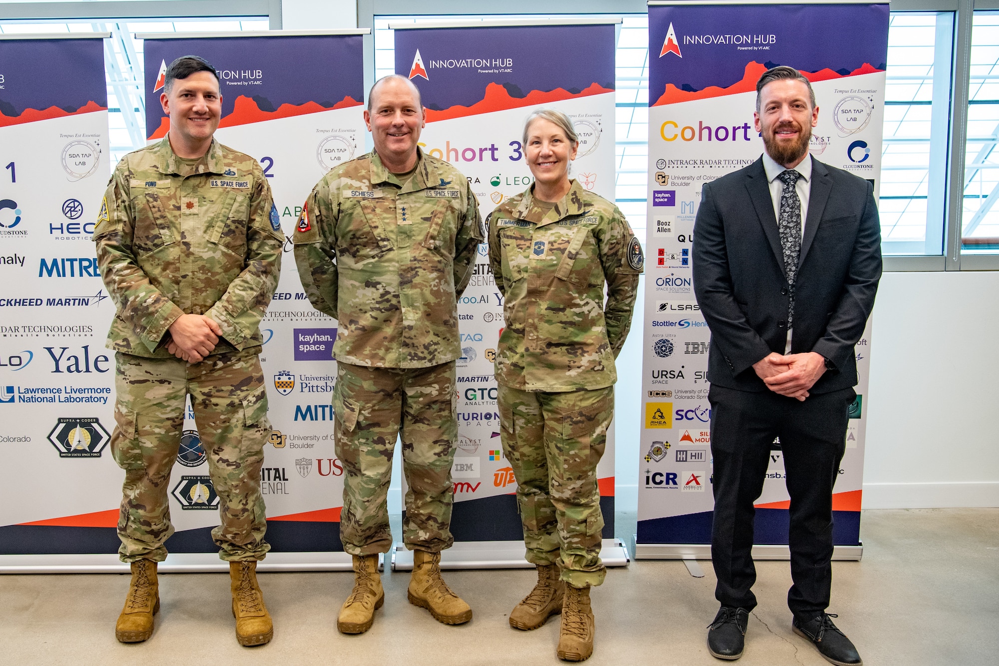 Three military members in uniform and man in suit pose for a photo in a row