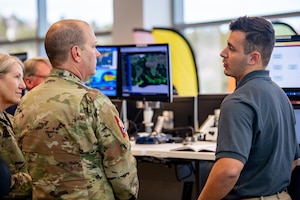 Man in military uniform listens to briefing from another man in polo shirt
