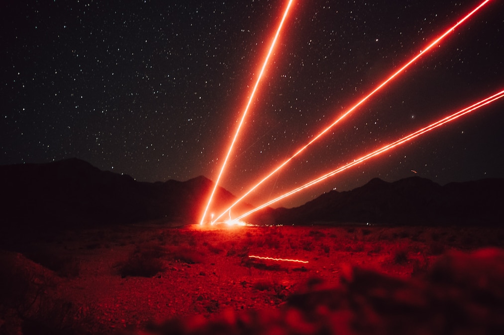 Three red beams of light blast from behind rocks toward a starry night sky in front of dark mountains.