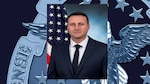 Photo of a white male in front of the America flag. He is wearing a suite.
