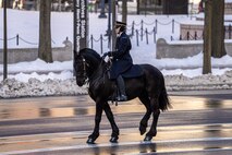 A soldier is riding a black horse down a street. There is snow on the ground in the distance.