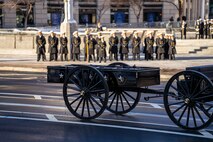 An empty caisson is parked on a street with Naval Academy cadets in the distance standing at parade rest.