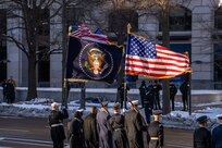 Service members are standing on the side of a street carrying the US and presidential flags, which are blowing in the wind.