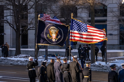 Service members are standing on the side of a street carrying the US and presidential flags, which are blowing in the wind.