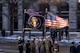 Service members are standing on the side of a street carrying the US and presidential flags, which are blowing in the wind.