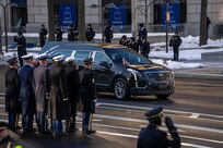 A hearse is driving down the street while uniformed service members line the street and some are saluting.
