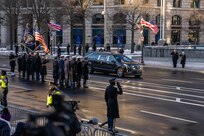 A hearse is driving down a street past ceremonial guard members carrying the US and presidential flags and other service members who are saluting.
