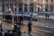 A hearse is driving down a street past ceremonial guard members carrying the US and presidential flags and other service members who are saluting.