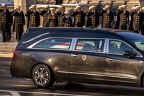 A hearse with the presidential seal on the door and a flag-draped coffin inside is driving past Navy cadets in dark overcoats and white hats who are saluting.