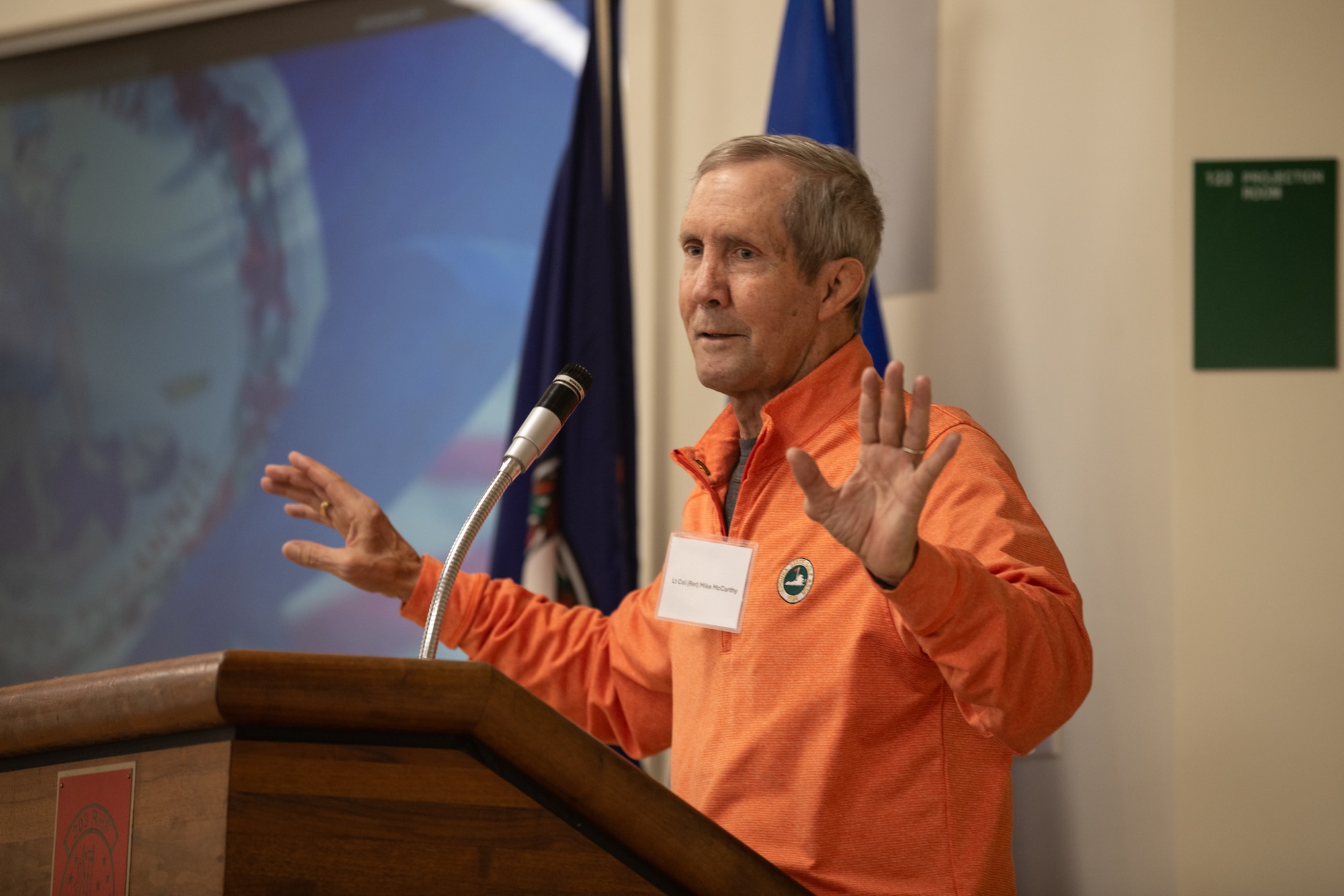 Man in orange shirt standing at podium with hands raised.