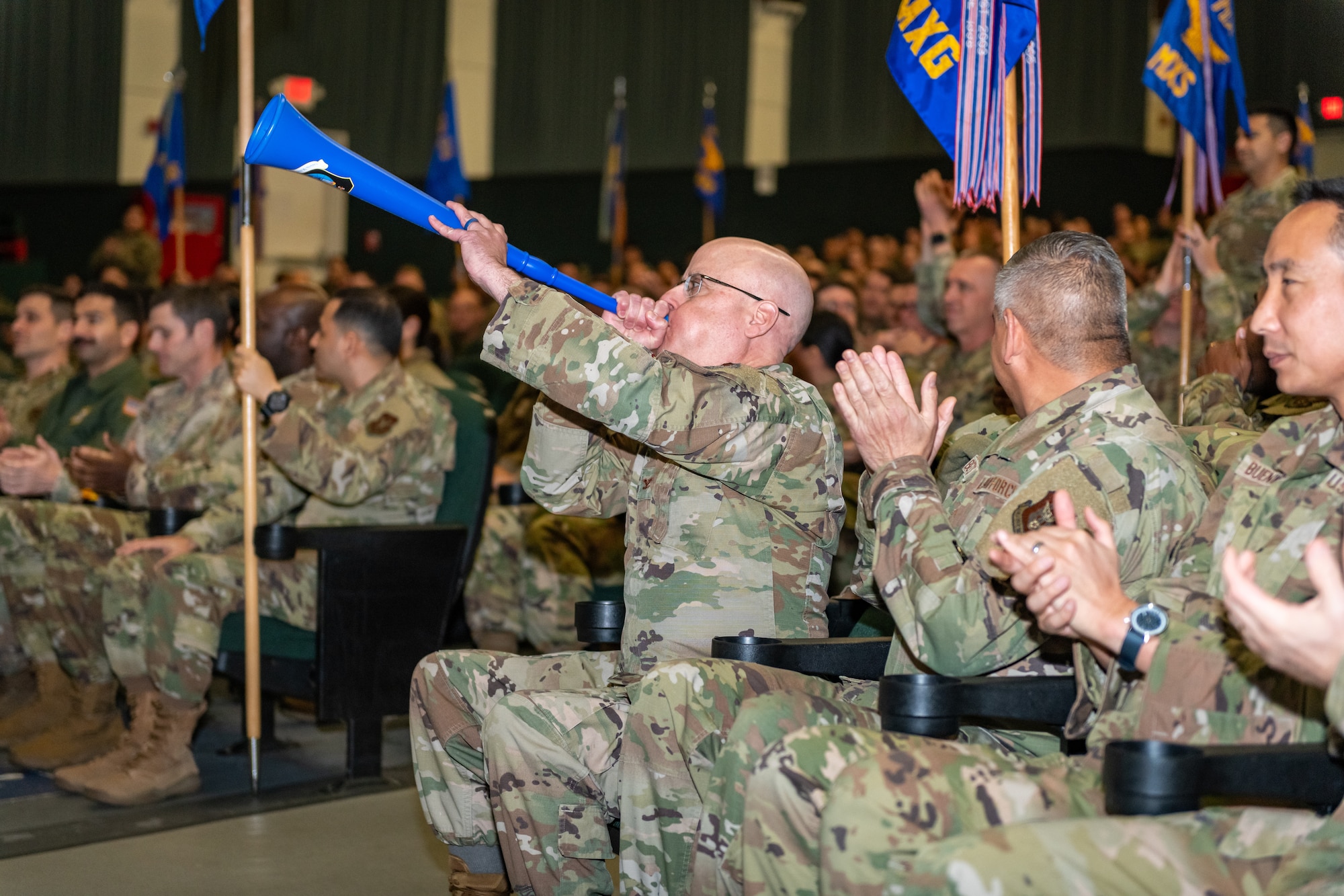 A military member blows into a horn in celebration surrounded by a crowd of cheering military members.