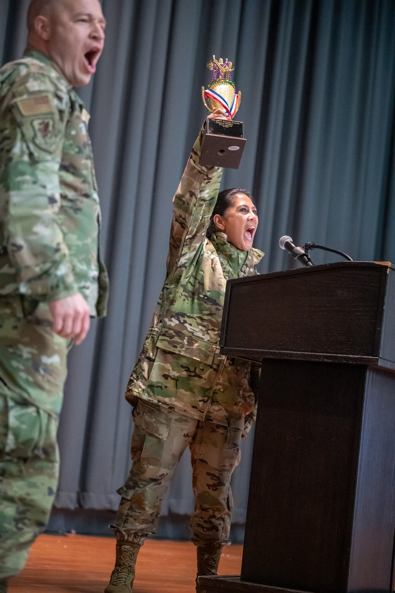 Two military members cheer loudly on the stage. One holding a trophy high about her head.