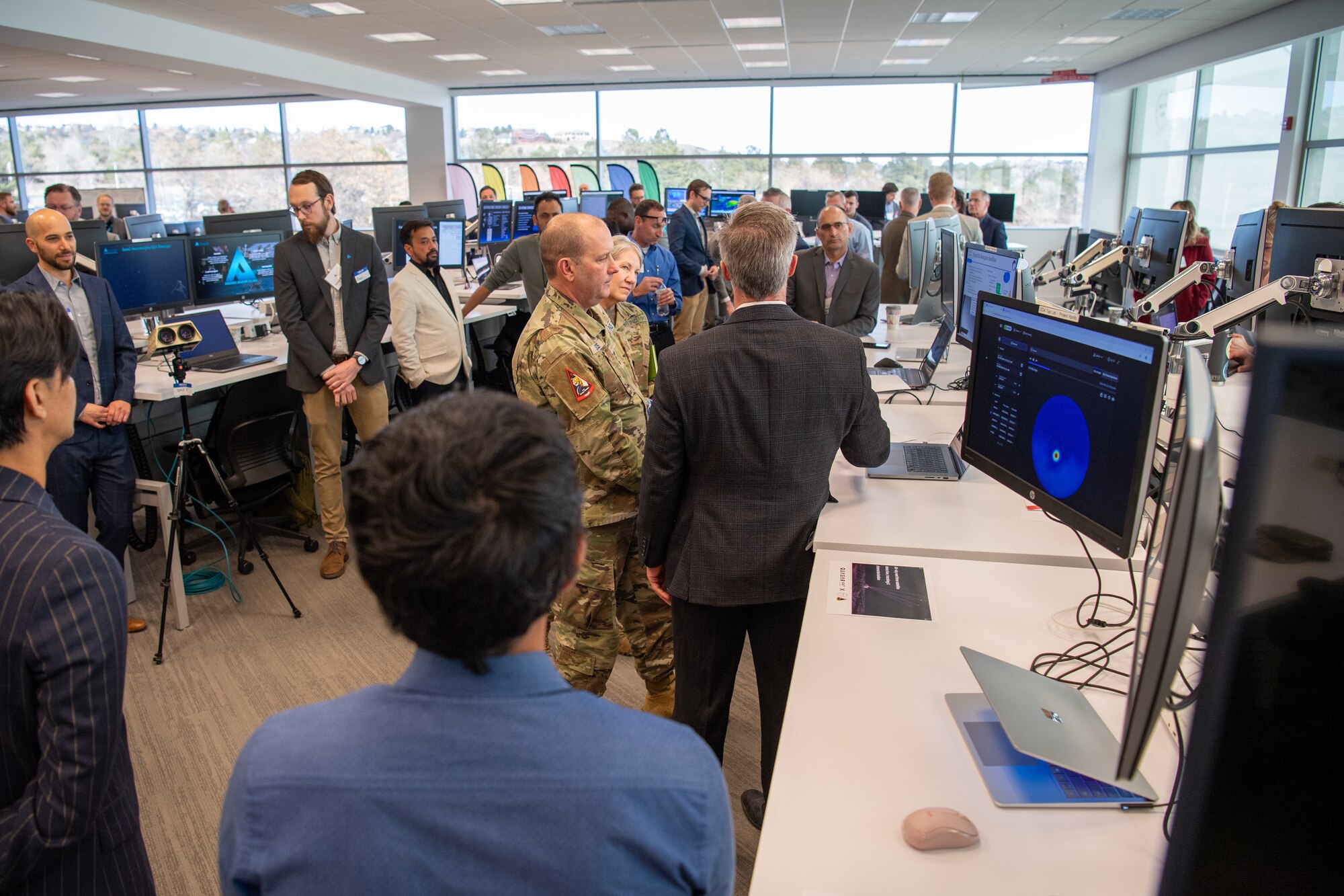 Military members listen to a brief in a room full of people and computers
