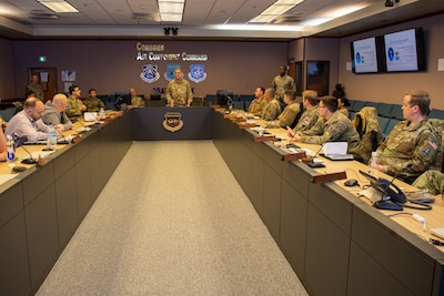 A dozen Space Force members sit around a U-shaped table, with a man standing at the head in the center of the photo and the words "Combined Air Component Command" on the wall behind him.